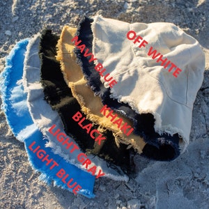 May include: A stack of six bucket hats in different colors, including off white, navy blue, khaki, black, light gray, and light blue. The hats have a frayed edge.