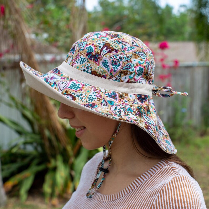 May include: A wide-brimmed sun hat with a paisley print in shades of brown, pink, teal, and white. The hat has a beige band and a tie at the back.