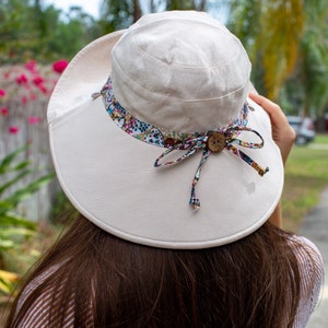 May include: A wide-brimmed sun hat with a white canvas exterior and a colorful floral patterned ribbon trim. The hat has a button accent on the ribbon.