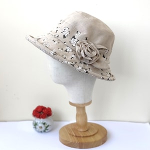 May include: A beige bucket hat with a floral pattern and a fabric rose. The hat is displayed on a mannequin head. The brim of the hat is decorated with small white and black flowers.