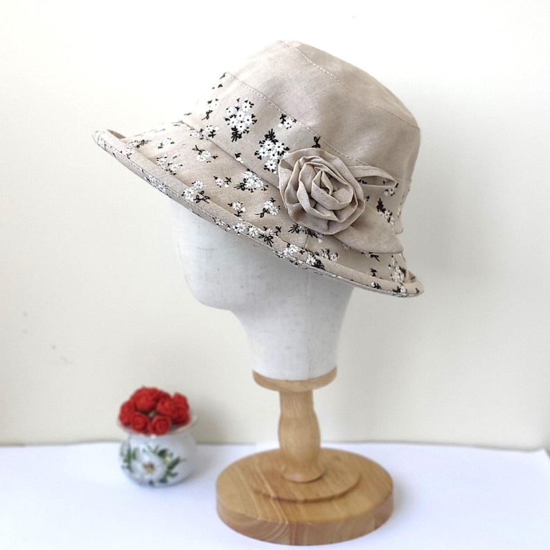 May include: A beige bucket hat with a floral pattern and a fabric rose. The hat is displayed on a mannequin head. The brim of the hat is decorated with small white and black flowers.