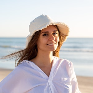 May include: A woman with long brown hair is wearing a white bucket hat and a white button-down shirt. She is standing in front of a body of water.