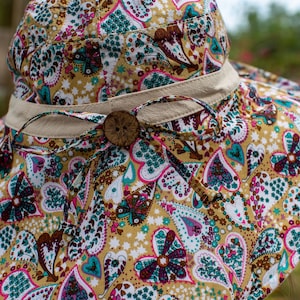 May include: A wide-brimmed sun hat with a brown button and a beige ribbon. The hat is made of a fabric with a paisley pattern featuring hearts, flowers, and stars in shades of pink, teal, brown, and white on a yellow background.