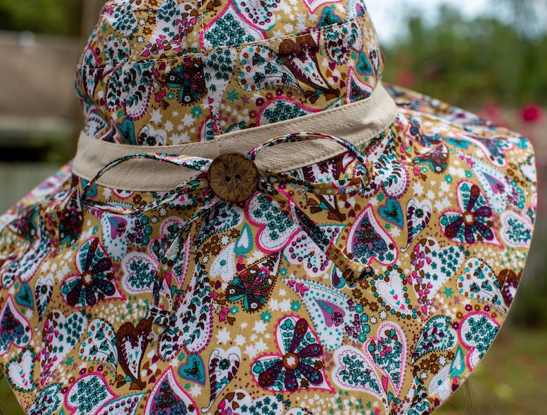 May include: A wide-brimmed sun hat with a brown button and a beige ribbon. The hat is made of a fabric with a paisley pattern featuring hearts, flowers, and stars in shades of pink, teal, brown, and white on a yellow background.