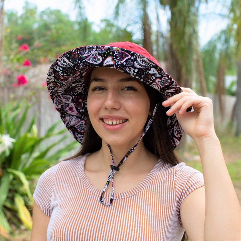 May include: A woman wearing a red and black patterned sun hat with a wide brim. The hat has a black chin strap and a black and white patterned lining. The woman is smiling and looking at the camera.