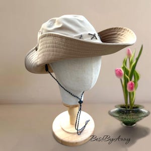 K&ouml;nnte beinhalten: Ein breitkrempiger Sonnenhut in hellem Beige mit einer dunkleren, hellbraunen Unterseite. Der Hut verf&uuml;gt &uuml;ber dekorative Kreuzstiche entlang der Krone und ein d&uuml;nnes, verstellbares Kinnband mit schwarz-wei&szlig;em Muster und einem Kunststoff-Stopper.