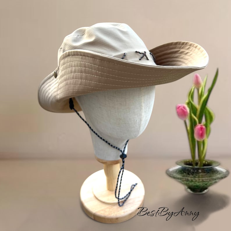 K&ouml;nnte beinhalten: Ein breitkrempiger Sonnenhut in hellem Beige mit einer dunkleren, hellbraunen Unterseite. Der Hut verf&uuml;gt &uuml;ber dekorative Kreuzstiche entlang der Krone und ein d&uuml;nnes, verstellbares Kinnband mit schwarz-wei&szlig;em Muster und einem Kunststoff-Stopper.