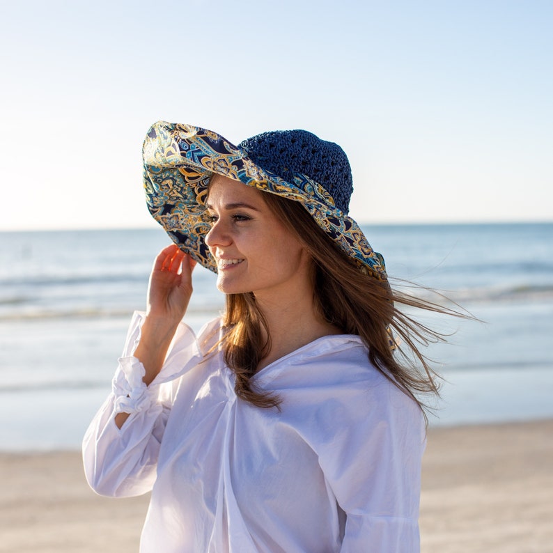 K&ouml;nnte beinhalten: Eine Frau tr&auml;gt ein wei&szlig;es Hemd und einen blauen und gelben gemusterten Sonnenhut mit blauem Rand. Sie steht an einem Strand mit dem Meer im Hintergrund.