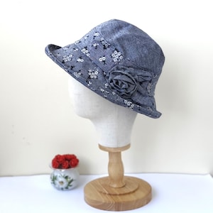 May include: A denim-style cloche hat with a floral pattern and a fabric rose accent. The hat is displayed on a mannequin head. The brim is decorated with small white flowers. The hat is approximately 4 inches in height.