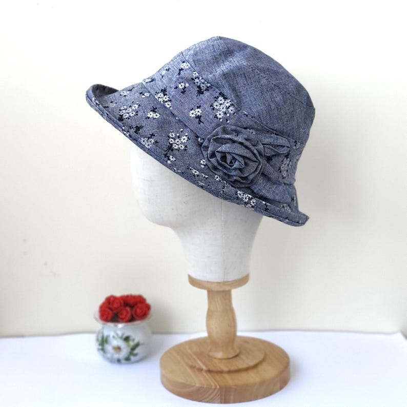 May include: A denim-style cloche hat with a floral pattern and a fabric rose accent. The hat is displayed on a mannequin head. The brim is decorated with small white flowers. The hat is approximately 4 inches in height.