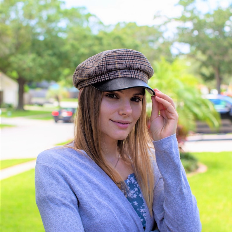 May include: A woman wearing a brown and black plaid newsboy cap with a black leather brim. She is wearing a light blue cardigan and has long brown hair.