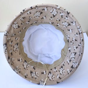 May include: A beige bucket hat with a floral pattern of small white flowers and black stems. The hat has a white interior and a beige brim. A white string is attached to the inside of the hat.