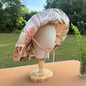 May include: A wide-brimmed sun hat with a floral print in shades of pink, brown, and blue. The hat has a pink ribbon tie.