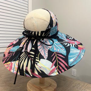May include: A wide-brimmed sun hat with a black base and a tropical leaf print in shades of pink, blue, yellow, and white. The hat has a black ribbon tied around the crown.