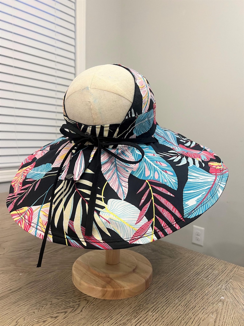 May include: A wide-brimmed sun hat with a black base and a tropical leaf print in shades of pink, blue, yellow, and white. The hat has a black ribbon tied around the crown.