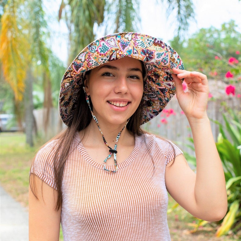 Pode incluir: Uma mulher vestindo uma camisa listrada marrom e branca com um chap&eacute;u de sol de aba larga com um padr&atilde;o paisley colorido. O chap&eacute;u tem uma correia de queixo.
