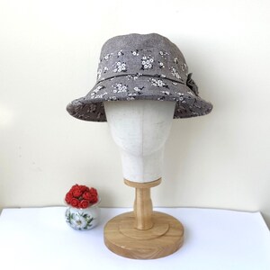 May include: A gray bucket hat with a white floral pattern is displayed on a mannequin head. The hat has a small bow on the side. The mannequin is on a wooden stand. A small ceramic pot with red roses is in the foreground.