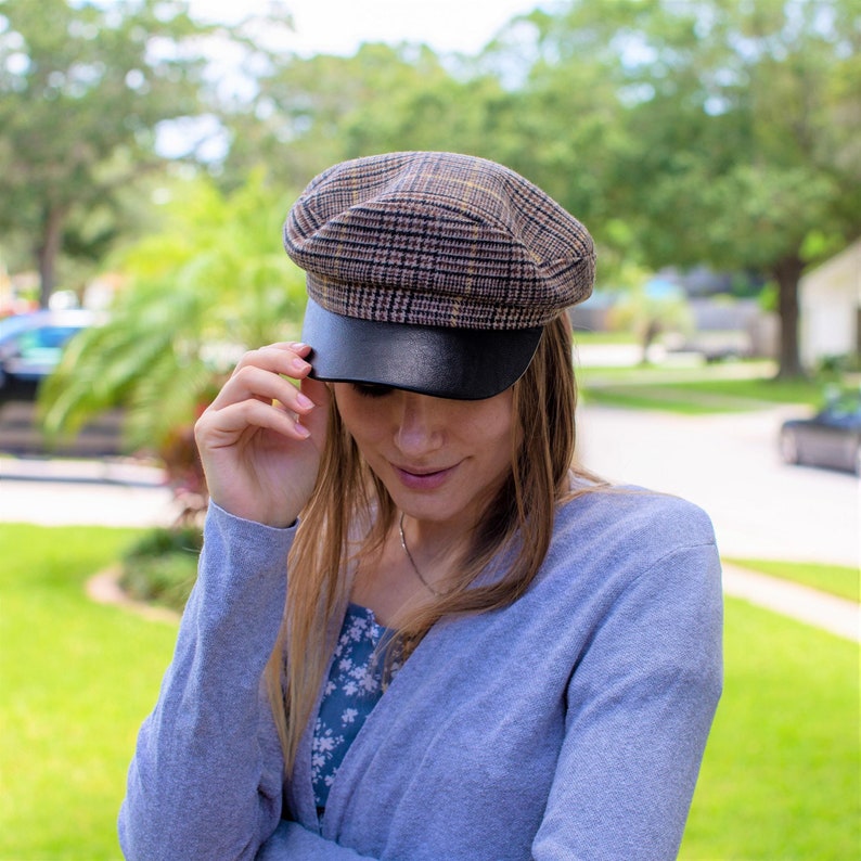 May include: A woman wearing a gray cardigan with a plaid and black newsboy cap. She is looking down at the camera.