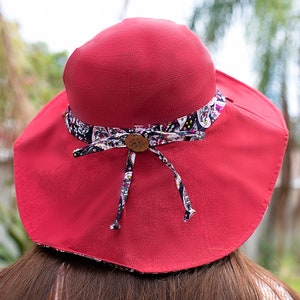 Pode incluir: Um chap&eacute;u de sol vermelho com aba larga e uma faixa de tecido estampada. A faixa &eacute; amarrada em um la&ccedil;o na parte de tr&aacute;s do chap&eacute;u.