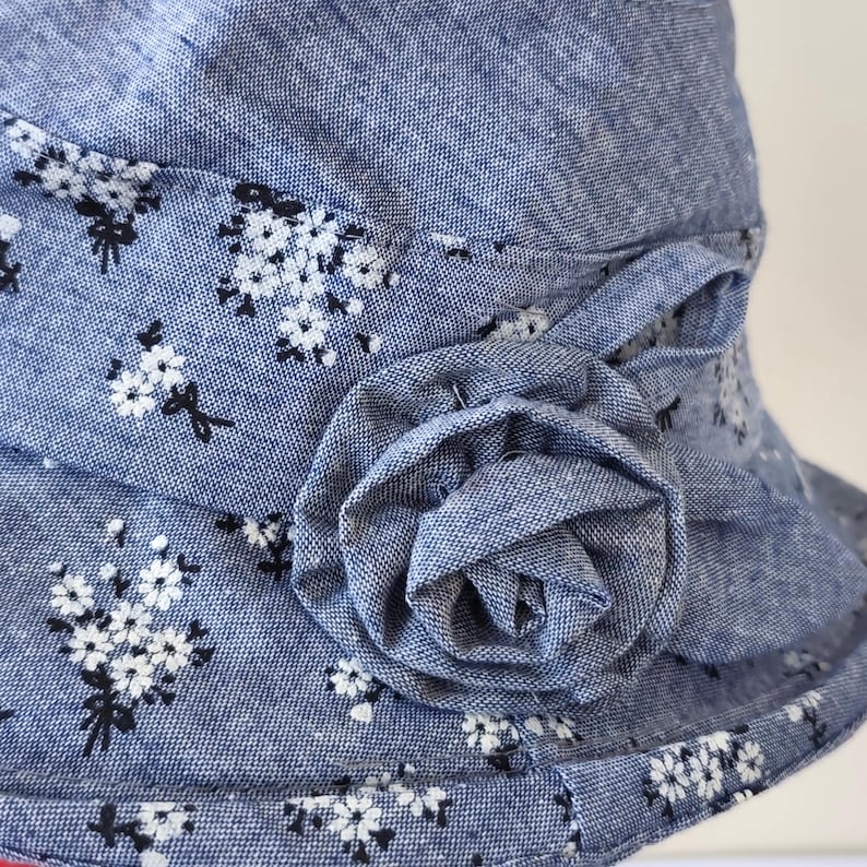 May include: A blue floral print hat with a decorative fabric flower. The hat features a white floral pattern on a blue background. The flower is made of the same fabric and is attached to the hat's band.