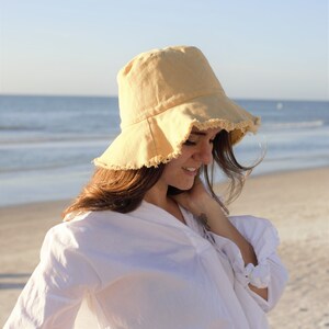 May include: A woman wearing a white shirt and a yellow bucket hat with frayed edges. She is standing on a sandy beach with the ocean in the background.