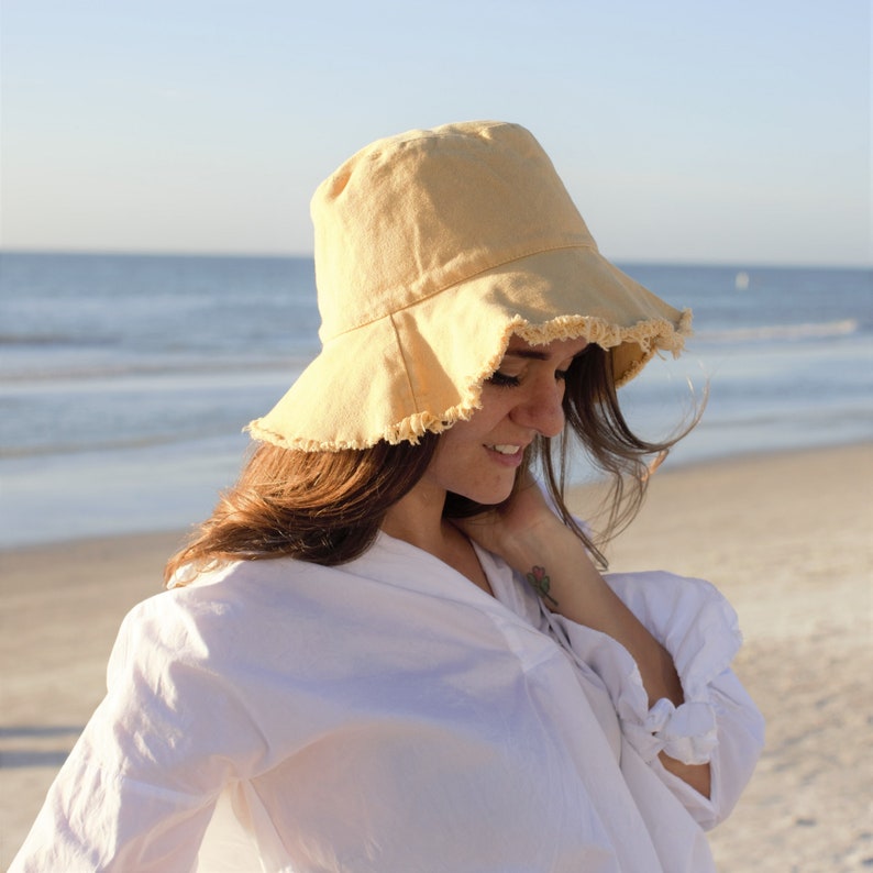May include: A woman wearing a white shirt and a yellow bucket hat with frayed edges. She is standing on a sandy beach with the ocean in the background.