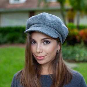 May include: A grey newsboy cap with a short peak. The cap is made of a soft, textured fabric. The hat is a classic style, suitable for various outfits. The cap is a neutral colour, making it versatile for different seasons.