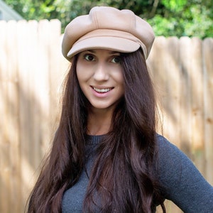 May include: A beige newsboy cap with a rounded crown and a small peak. The cap is made of a soft material. A woman with long, dark brown hair wears the cap and a grey sweater. A wooden fence forms the background.