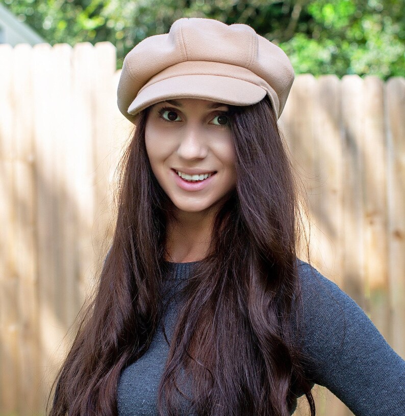 May include: A beige newsboy cap with a rounded crown and a small peak. The cap is made of a soft material. A woman with long, dark brown hair wears the cap and a grey sweater. A wooden fence forms the background.