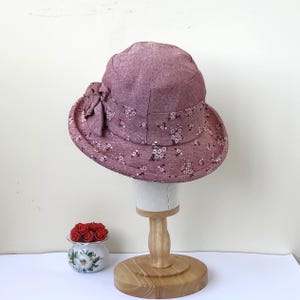 May include: A mauve-colored sun hat with a floral pattern and a decorative bow. The hat is displayed on a wooden stand. The brim and crown are adorned with small white flowers. The hat is approximately 10 inches in diameter.