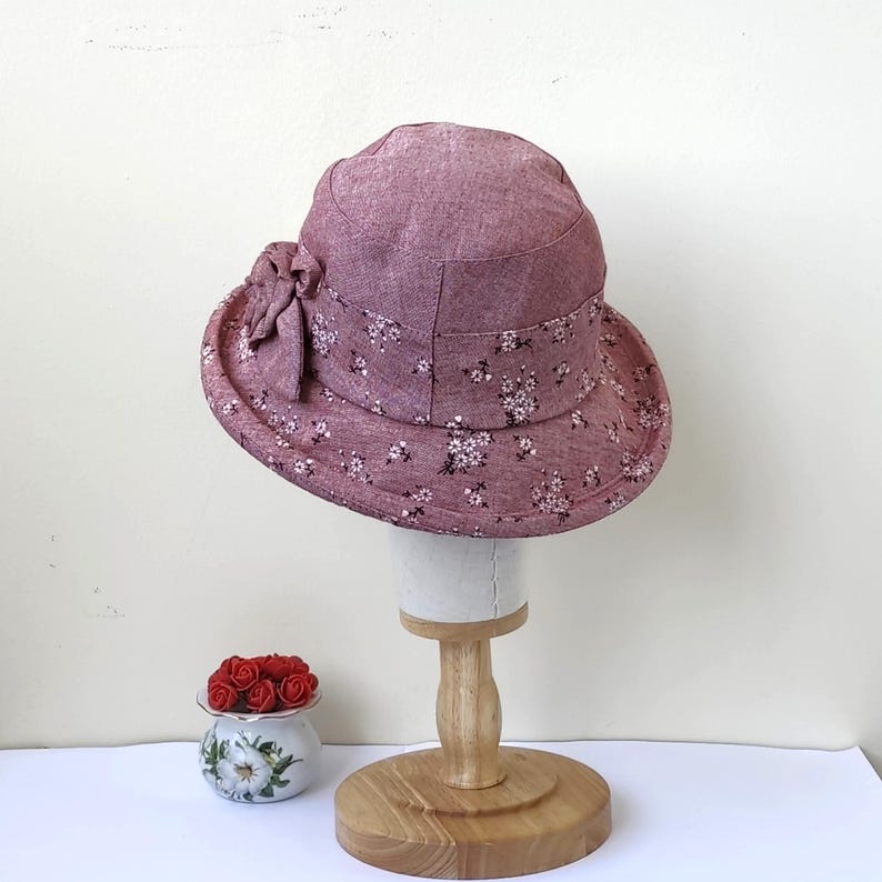 May include: A mauve-colored sun hat with a floral pattern and a decorative bow. The hat is displayed on a wooden stand. The brim and crown are adorned with small white flowers. The hat is approximately 10 inches in diameter.