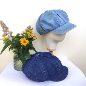 May include: Two denim newsboy caps in shades of blue. One is light blue, the other is indigo. The lighter blue cap is on a mannequin head. A vase of flowers is to the left of the caps. The caps are a stylish accessory.