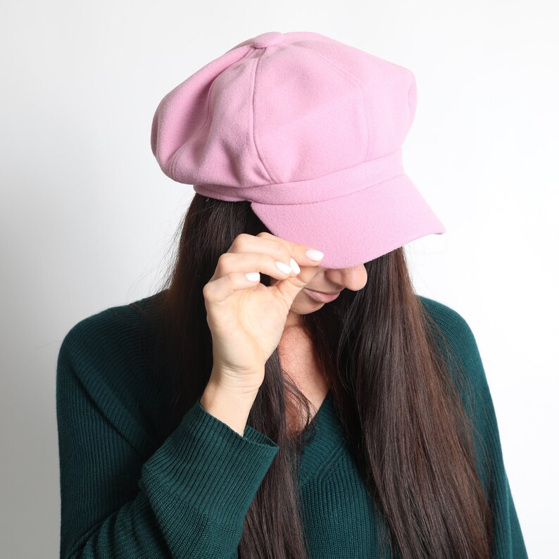 May include: A pale pink newsboy cap with a rounded crown and a short, stiff peak. The cap is made of a soft, textured fabric. The person in the image is wearing a dark green jumper.