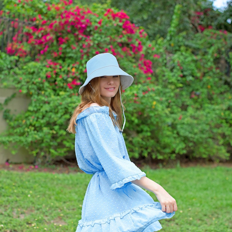 May include: A woman wearing a light blue dress with a light blue bucket hat. The dress has a ruffled hem and a white polka dot pattern. She is standing in a grassy area with green foliage in the background.