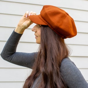 May include: A burnt orange newsboy cap with a rounded crown and a short, stiff peak. The cap is made of a soft, textured fabric. The person is wearing a grey long-sleeved top.