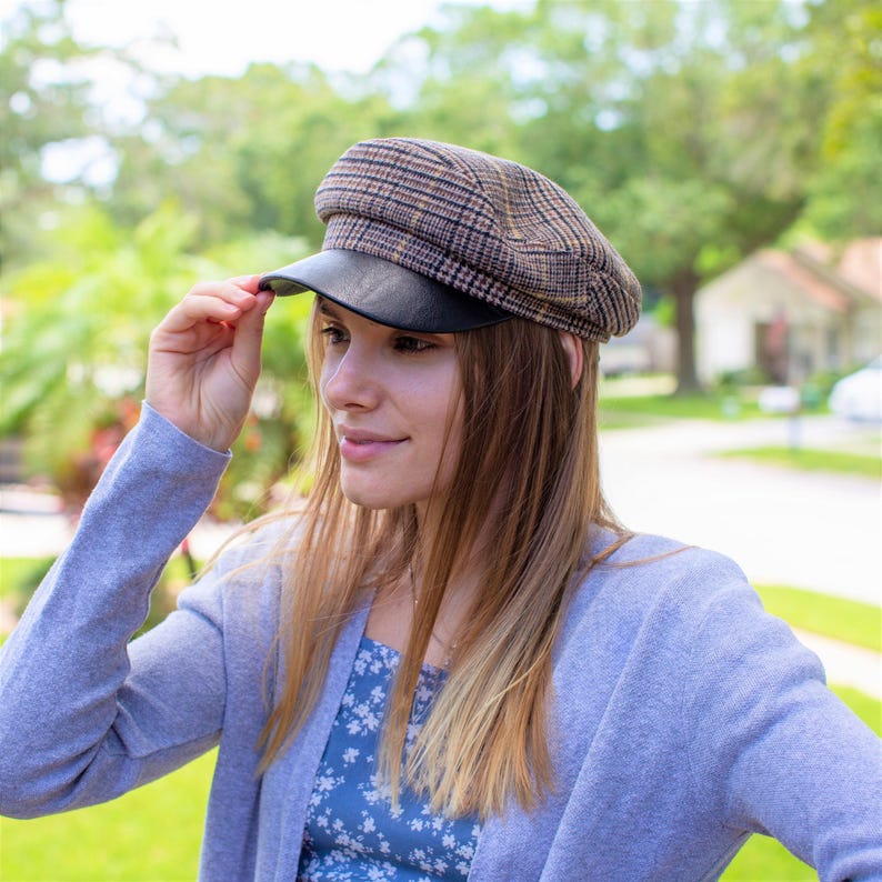 May include: A woman wearing a light blue cardigan with a floral print top underneath. She is wearing a brown and black plaid newsboy cap with a black leather brim.