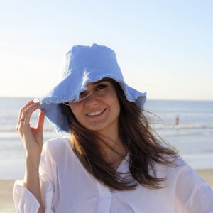 May include: A woman wearing a light blue bucket hat with frayed edges and a white shirt. She is smiling and looking at the camera. The background is a beach with blue water and a clear sky.