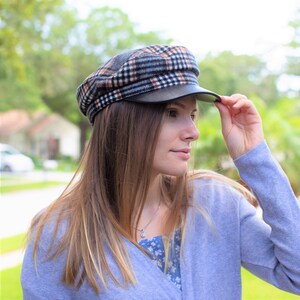 May include: A woman wearing a gray sweater and a black and white plaid newsboy cap with a black leather brim. The cap has a slightly curved brim and is tilted slightly to the side.