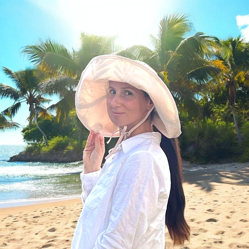 May include: A woman wearing a white shirt and a large, wide-brimmed sun hat with a light brown color. The hat has a long, flowing tie that is tied under her chin. She is standing on a sandy beach with palm trees in the background.