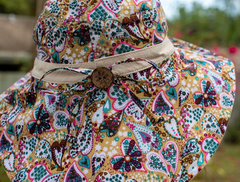Pode incluir: Um chap&eacute;u de sol de aba larga com estampa paisley em tons de marrom, rosa, azul-turquesa e branco. O chap&eacute;u tem uma fita bege amarrada na coroa com um bot&atilde;o de madeira.
