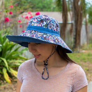Pode incluir: Uma mulher usando um chap&eacute;u de sol azul com padr&atilde;o floral de aba larga e uma correia de queixo. O chap&eacute;u tem uma faixa azul ao redor da coroa.