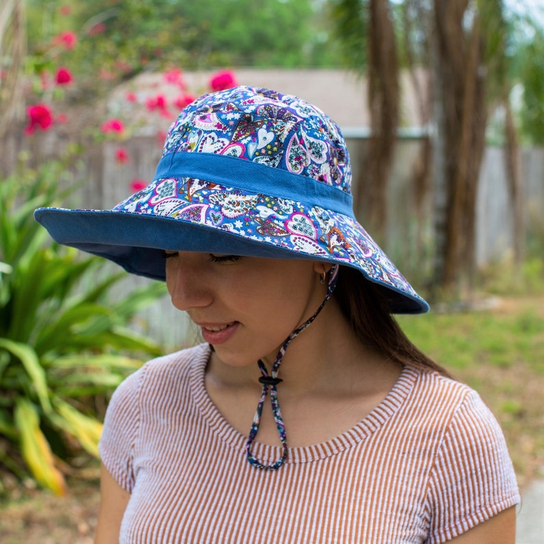 Pode incluir: Uma mulher usando um chap&eacute;u de sol azul com padr&atilde;o floral de aba larga e uma correia de queixo. O chap&eacute;u tem uma faixa azul ao redor da coroa.
