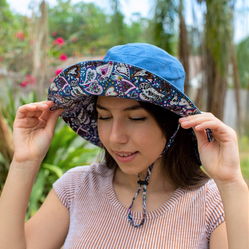 Pode incluir: Uma mulher usando um chap&eacute;u de sol azul com uma ampla aba e um padr&atilde;o floral. O chap&eacute;u tem uma correia de queixo e &eacute; amarrado na parte de tr&aacute;s do pesco&ccedil;o.