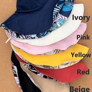 May include: A stack of eight sun hats in various colors, including navy, ivory, pink, yellow, red, beige, and black. Each hat has a different colored lining with a tropical leaf pattern.