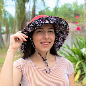 Pode incluir: Uma mulher usando um chap&eacute;u de sol de aba larga com padr&atilde;o vermelho e preto. O chap&eacute;u tem uma correia de queixo e &eacute; amarrado na parte de tr&aacute;s do pesco&ccedil;o. A mulher est&aacute; sorrindo e olhando para a c&acirc;mera.