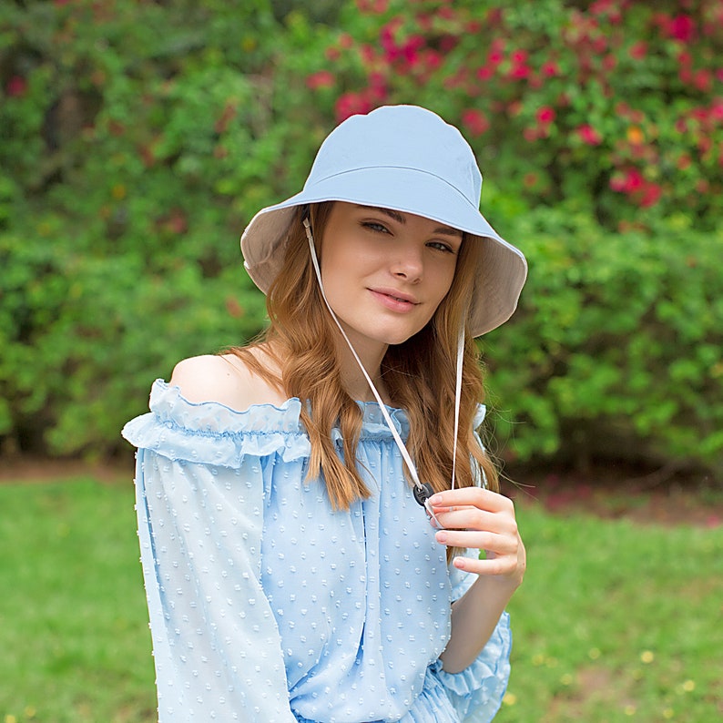 May include: A light blue bucket hat with a white chin strap. The hat is worn by a person with long, wavy, light brown hair. The person is wearing a light blue, off-the-shoulder blouse with white polka dots.