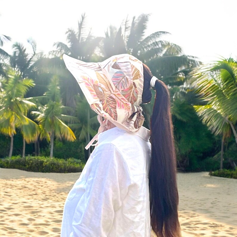 May include: A white sun hat with a floral print and a wide brim. The hat is worn by a person with long dark hair. The person is standing on a beach with palm trees in the background.