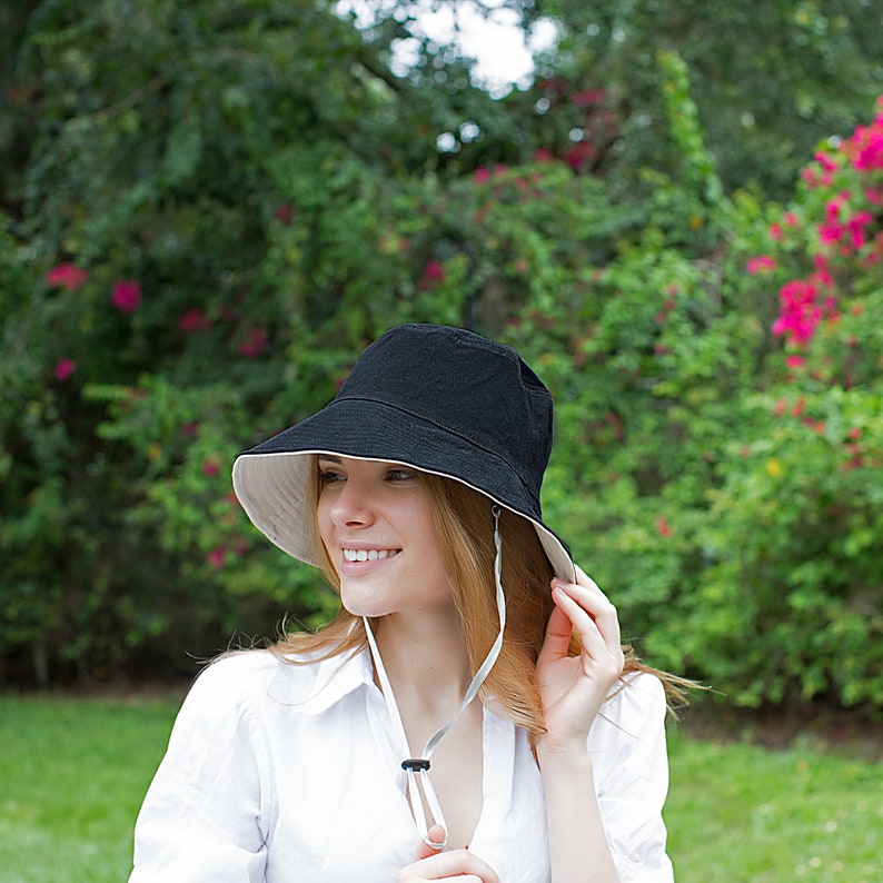 May include: A woman wearing a black and white bucket hat with a chin strap. The hat has a wide brim and is made of a lightweight material. The woman is smiling and looking to the side.