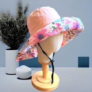 May include: A pink and floral sun hat with a black chin strap. The hat is on a wooden display stand.