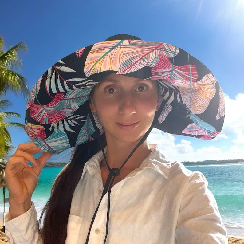 May include: A woman wearing a wide-brimmed sun hat with a black background and colorful tropical leaf print. The hat has a black ribbon tied under her chin.
